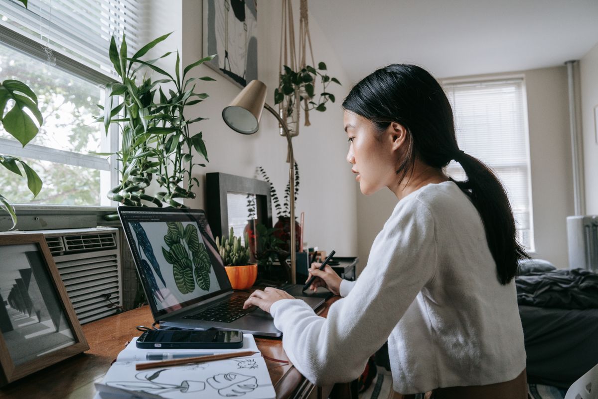 An Asian woman designer working on graphics on a laptop with a drawing tablet and sketches on the desk — the design stage before lorry sticker artwork is approved for print