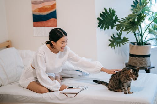 Woman in White Long Sleeves Sitting on White Bed with Her Tabby Cat