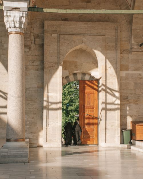 Free Two women exit a mosque courtyard through a large wooden door under sunlight. Stock Photo