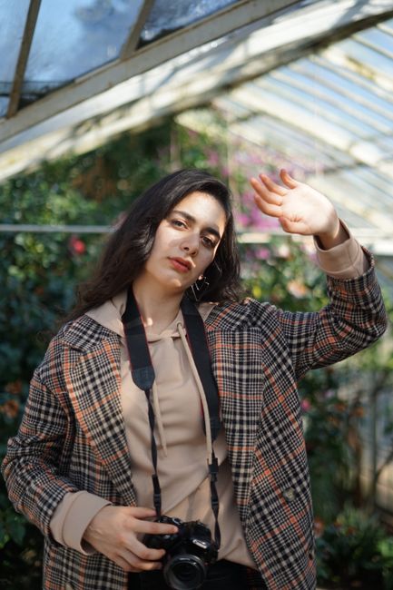 Portrait of a woman in a greenhouse holding a DSLR camera, sunlight filtering through glass.