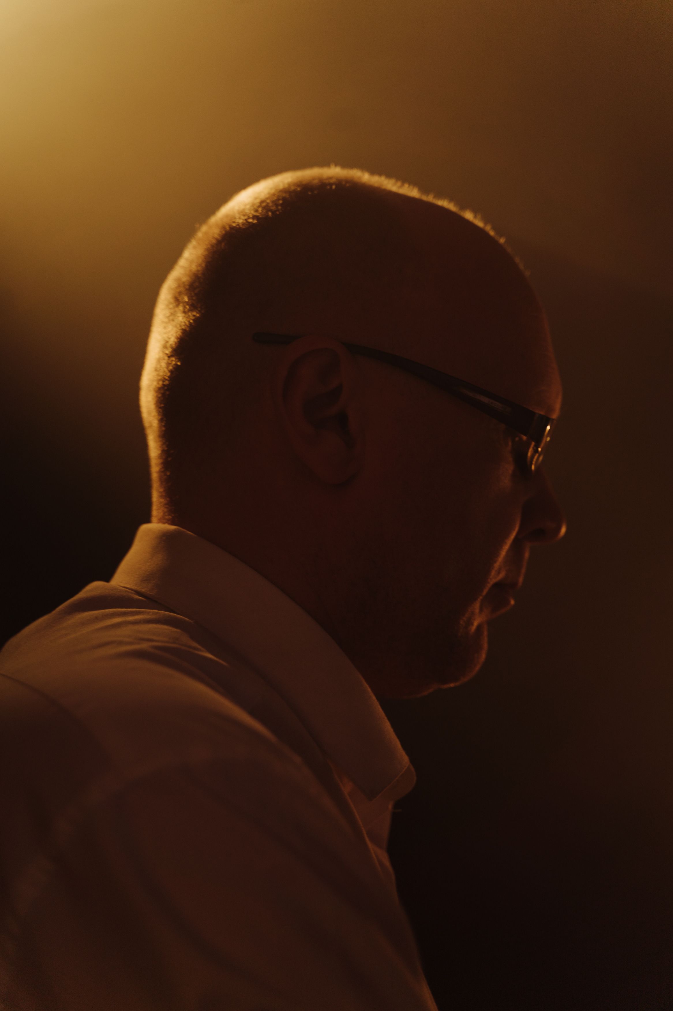 Side profile of a bald man in glasses, backlit with warm lighting, creating a dramatic effect.