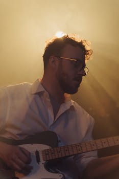 Musician playing guitar on stage, backlit by atmospheric stage lights.