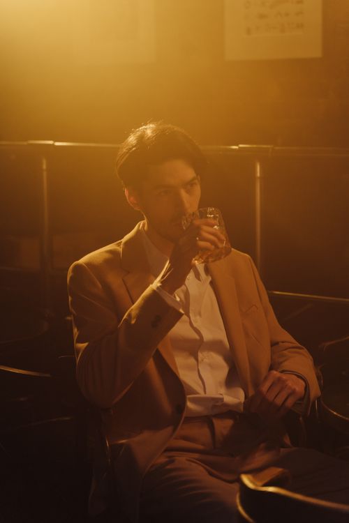 Free Sophisticated Asian man in a suit sipping whiskey in a dimly lit bar, exuding a warm and moody atmosphere. Stock Photo