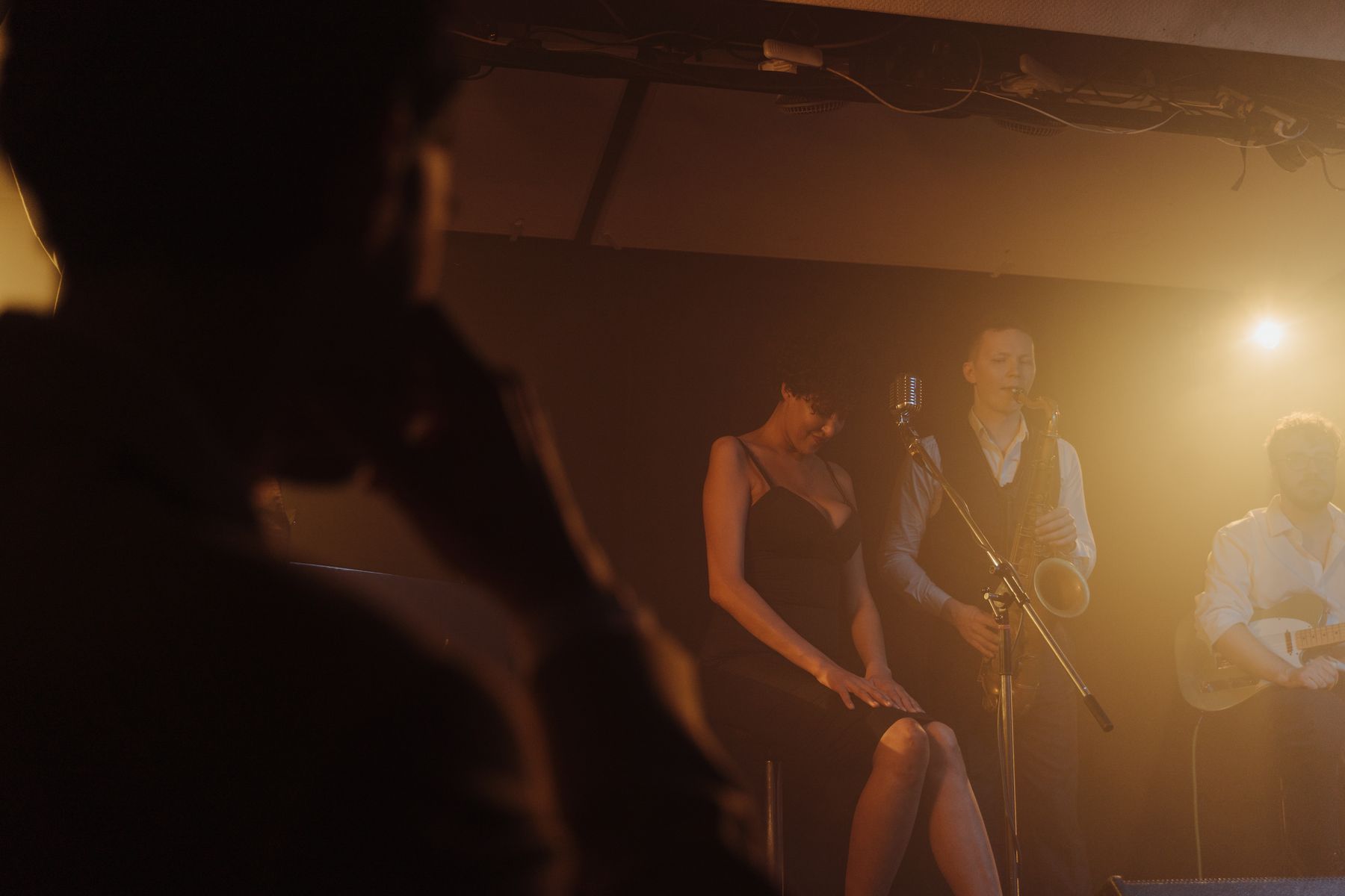 A saxophonist and singer performing together in a dimly lit room, warm light behind them.