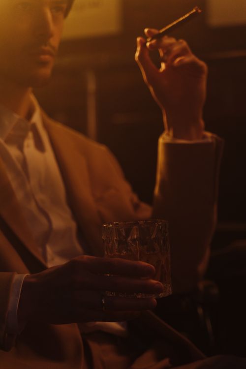 Free A contemplative man elegantly holding a whiskey glass and cigar in a warm, ambient setting. Stock Photo
