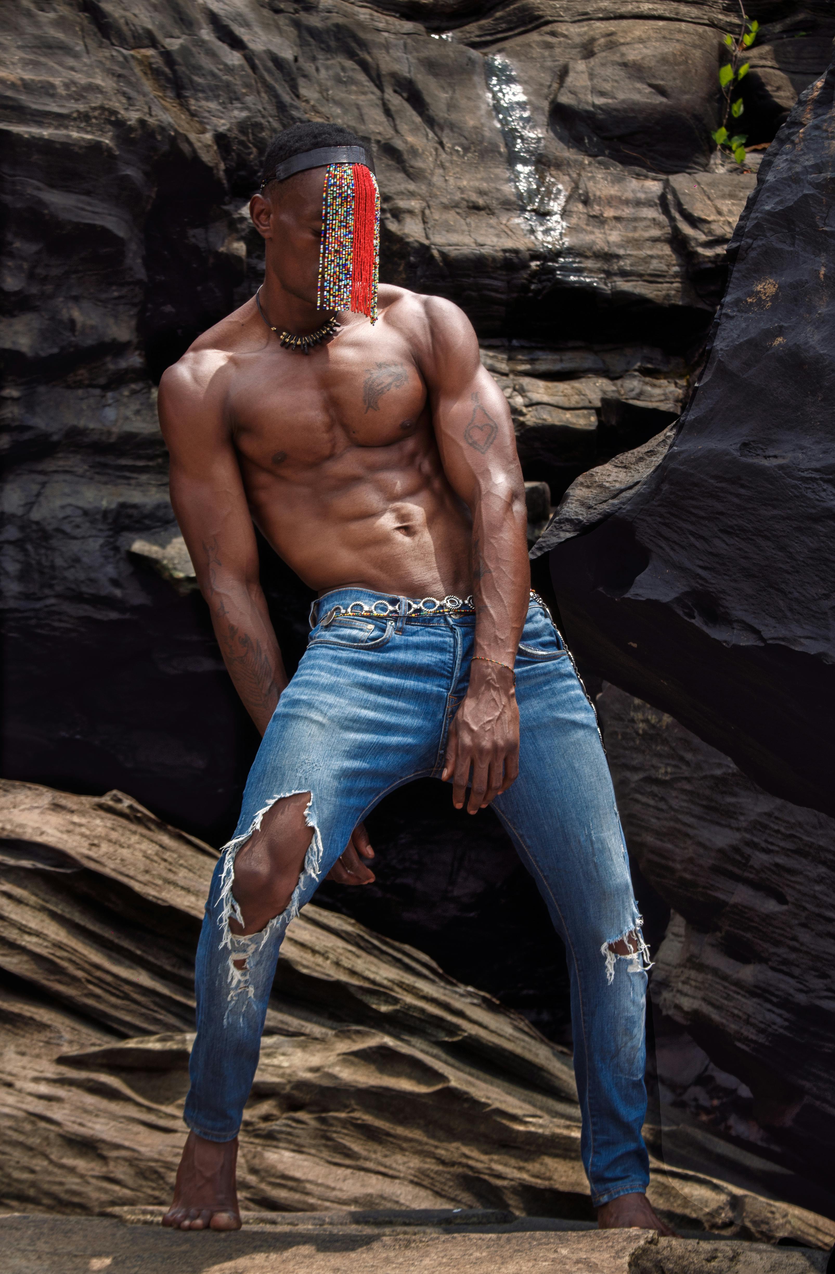 Shirtless man in ripped jeans posing confidently against rock formations outdoors.