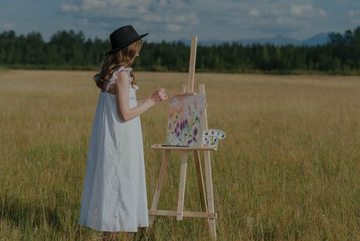 A woman paints a field landscape under a bright summer sky, capturing nature's beauty.