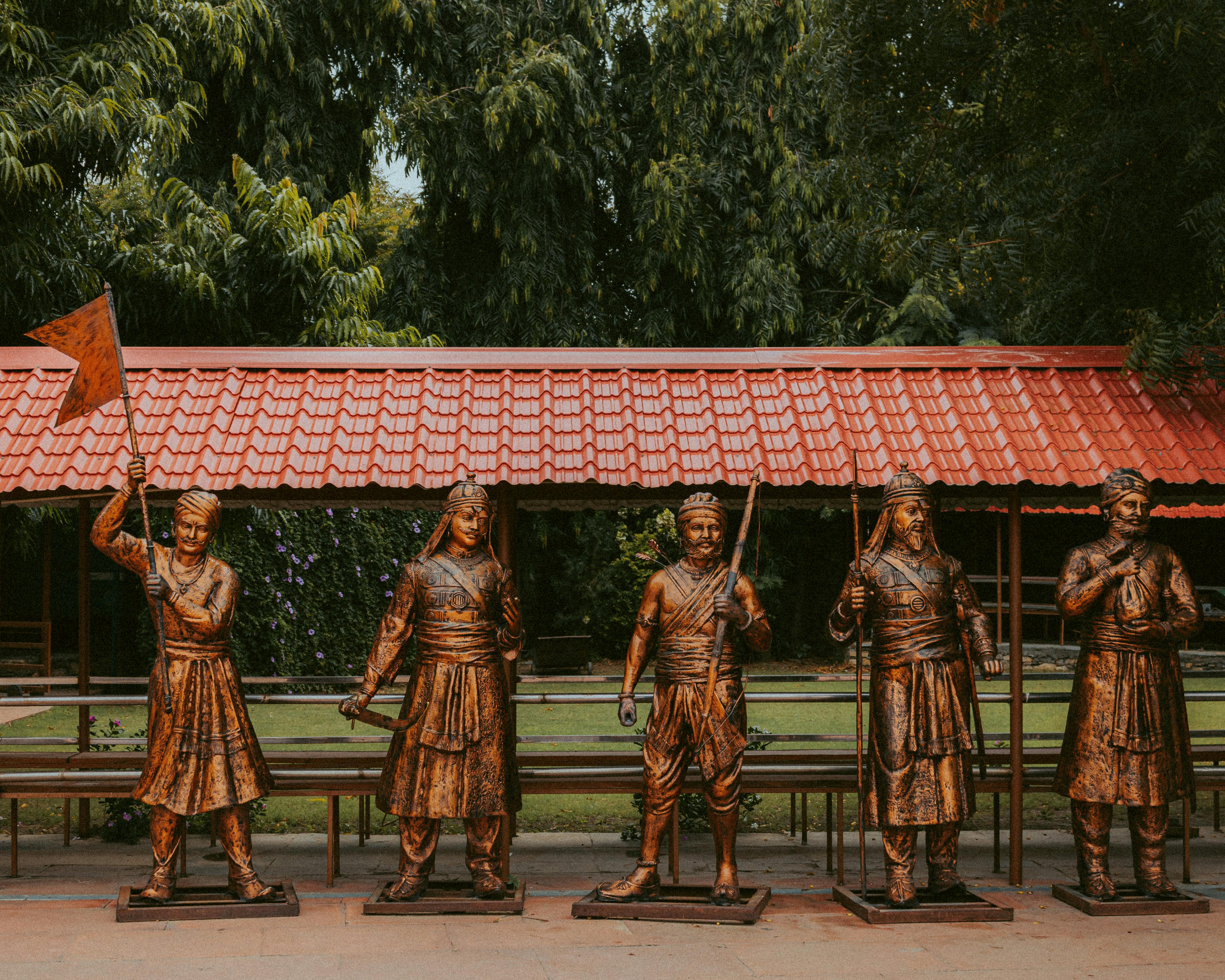 Copper-colored warrior statues under a red tiled roof amidst greenery.