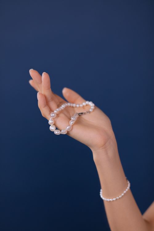 Free Elegant hand holding a pearl bracelet against a dark blue background, showcasing luxury and sophistication. Stock Photo