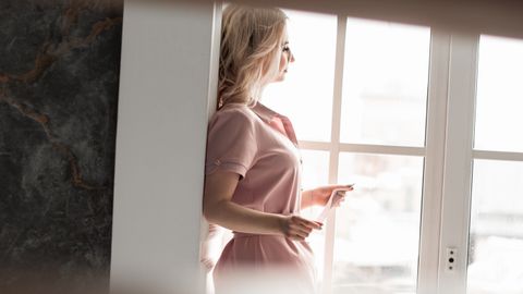 A woman in pink standing thoughtfully by a window. Soft natural light highlights her contemplative mood.