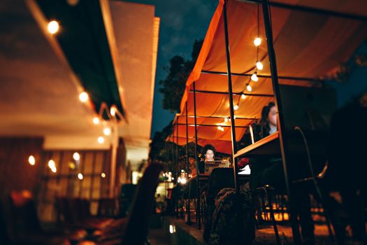 A vibrant evening cafe scene with people working on laptops under illuminated awnings.