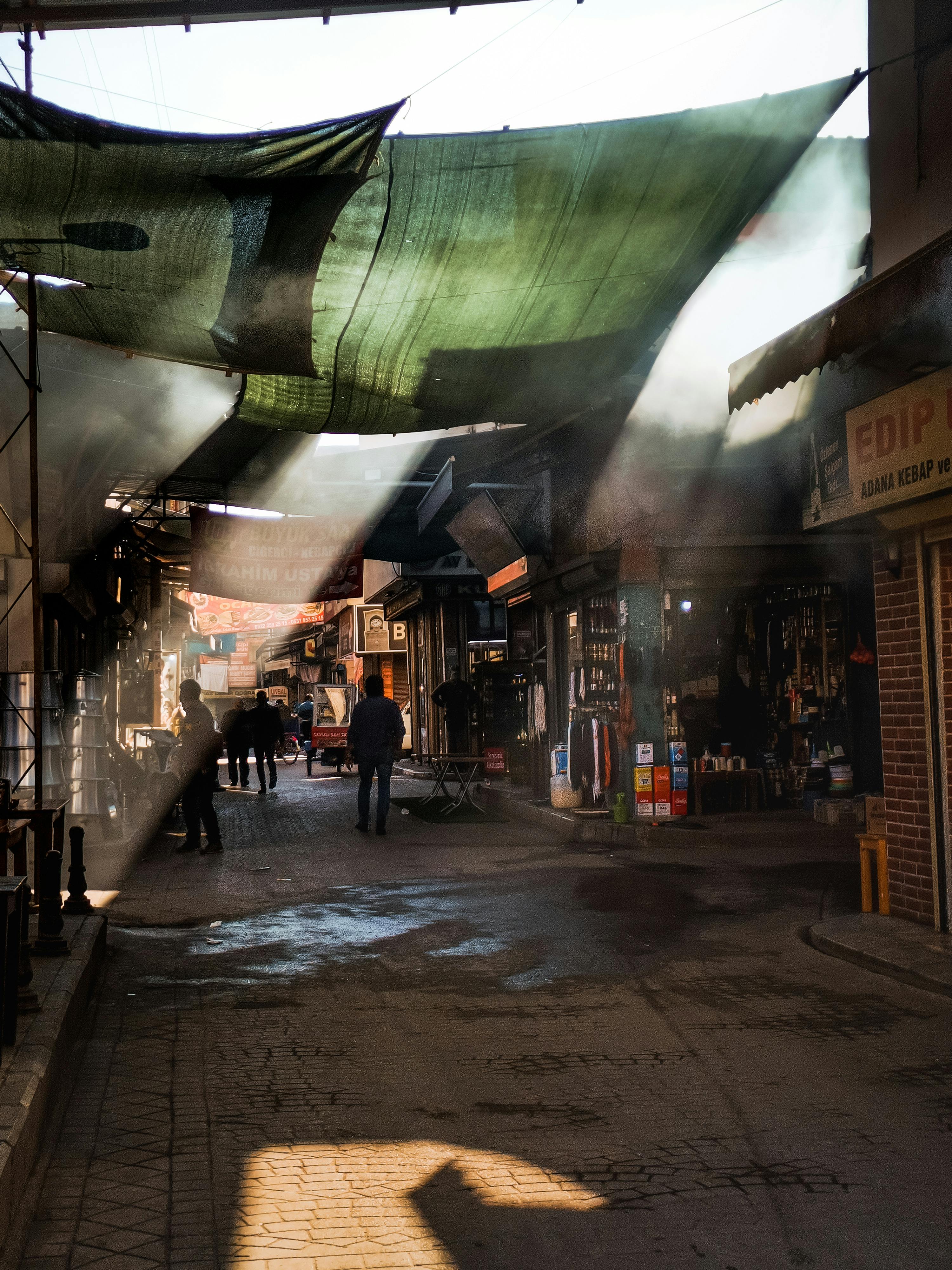 A vibrant marketplace with shops illuminated by dramatic sunlight beams through fabric canopies.
