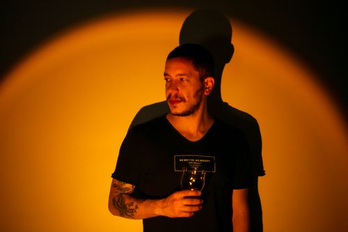 Free A man in a black shirt holds a glass against a warm light backdrop, casting a strong shadow. Stock Photo