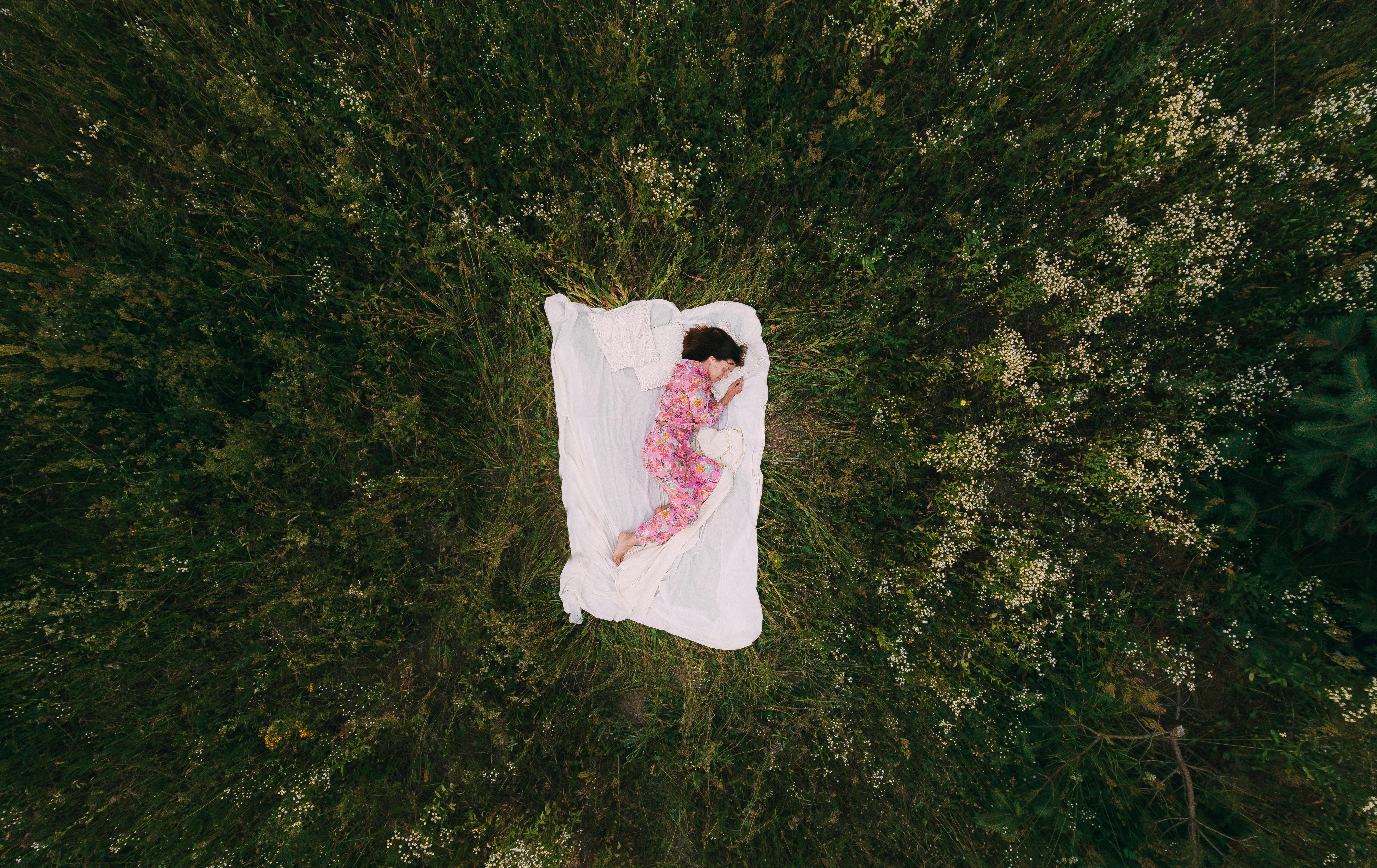 A woman in floral pajamas sleeps on a white blanket in a lush, green field viewed from above.