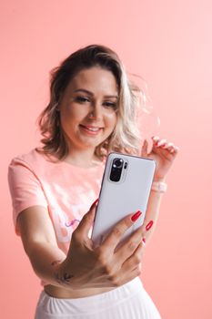 Smiling woman holds smartphone for a selfie against a pink background. Ideal for lifestyle and technology themes.