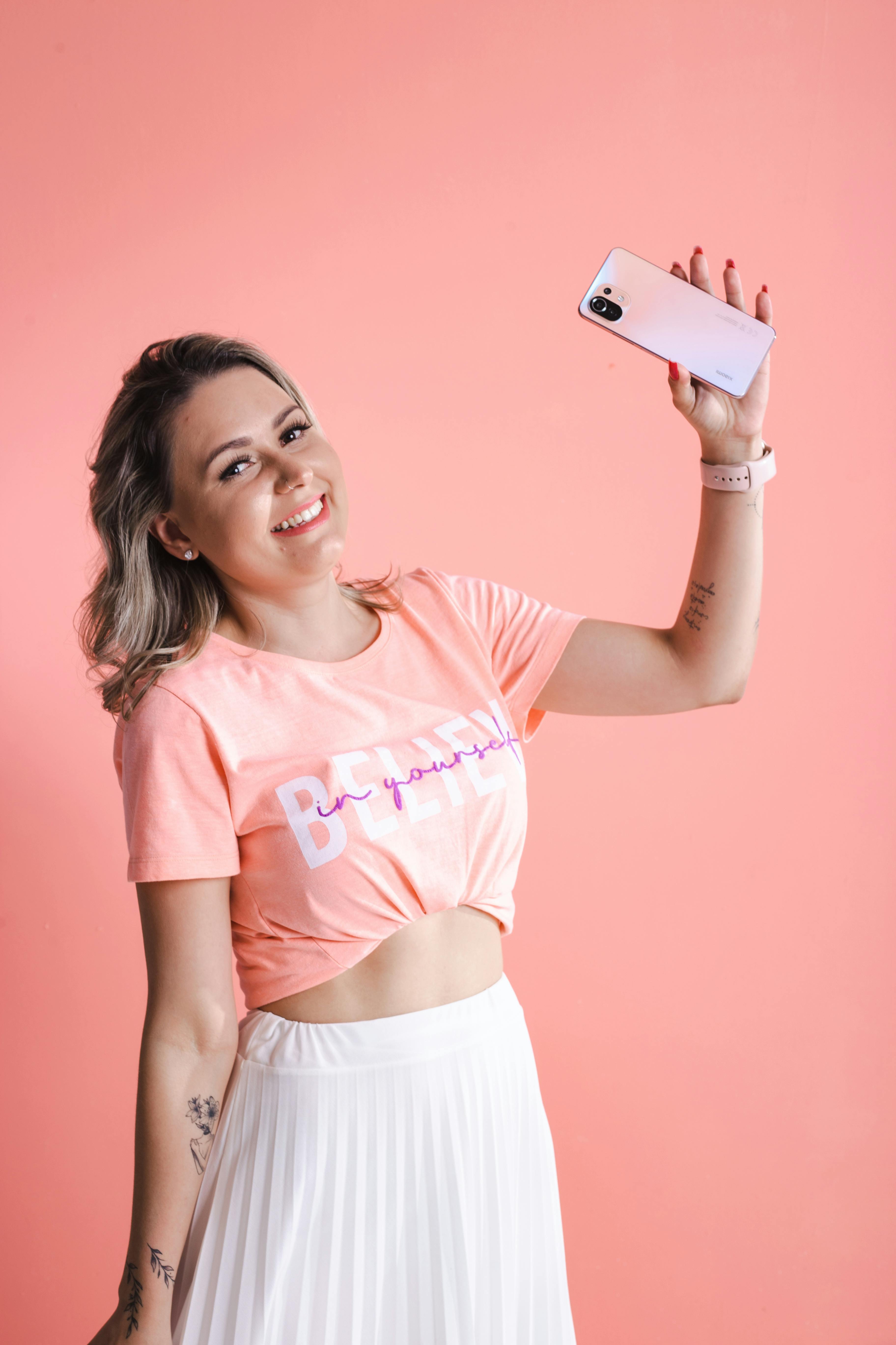 Young woman in pink outfit taking a joyful selfie on a pink background.