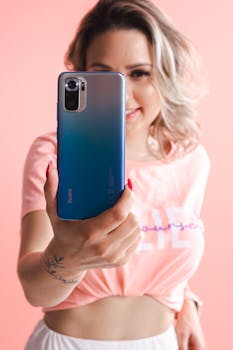 Woman smiling and posing with blue smartphone against pink studio background.