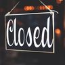 A black wooden sign with the word Closed written in white cursive, hanging in front of warm, blurred bokeh lights.