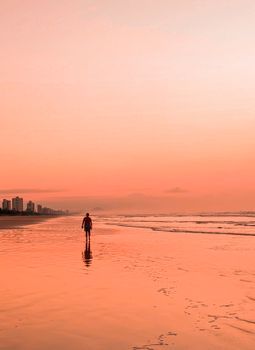 A silhouetted person walks along the beach reflecting the vivid orange sky during sunset.