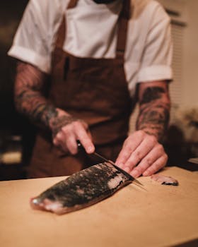 Tattooed chef skillfully slices salmon in kitchen setting.