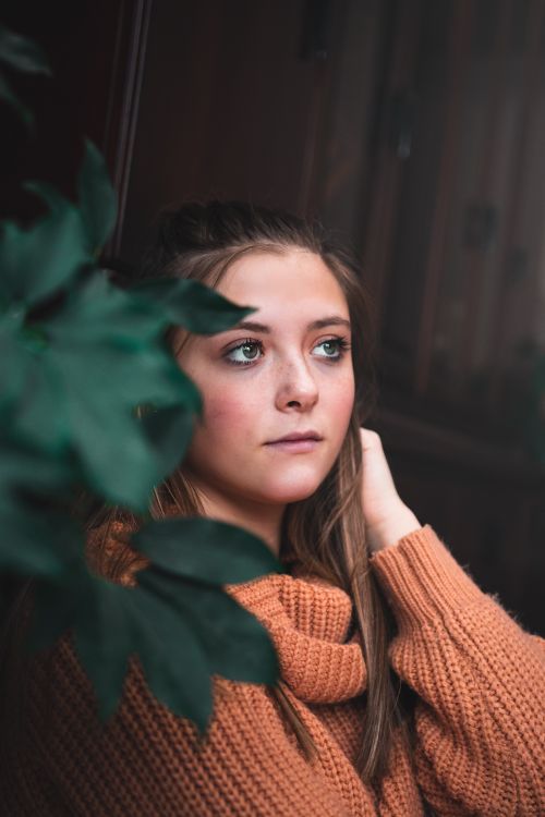 Free A young woman in a cozy knitted sweater gazes thoughtfully, framed by green indoor plants. Stock Photo