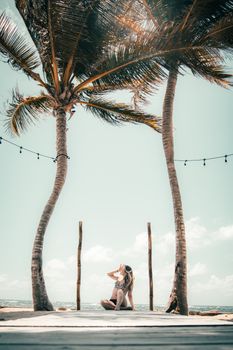 A woman sitting under swaying coconut trees on a tropical beach, basking in the sun.