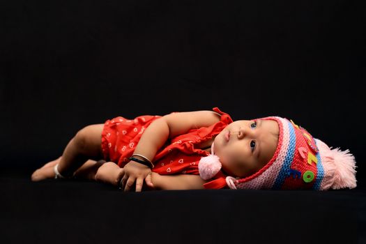 Cute baby girl wearing a colorful knit hat, lying on a dark background indoors.