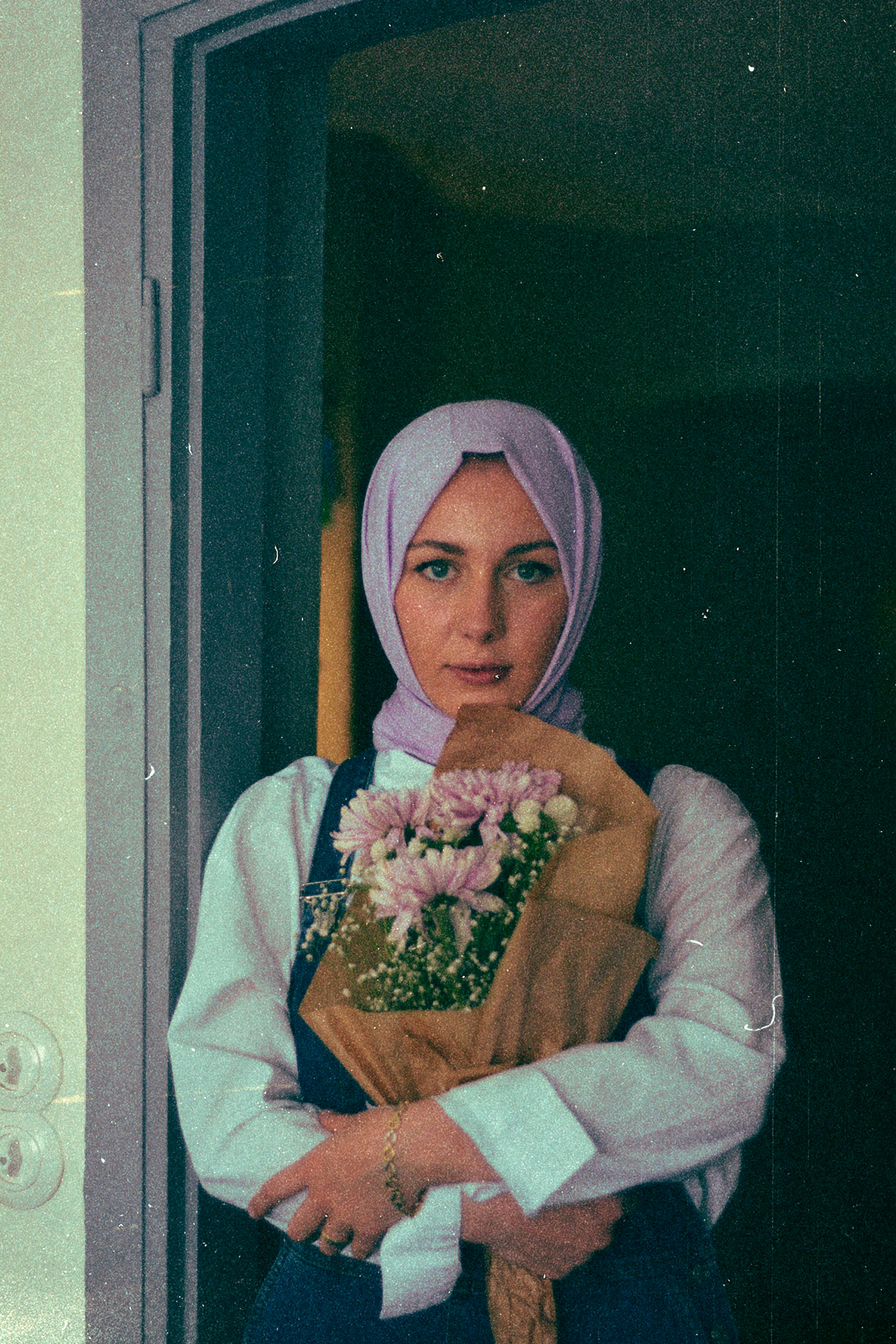 A woman in a hijab holding a bouquet of flowers, standing in a doorway.