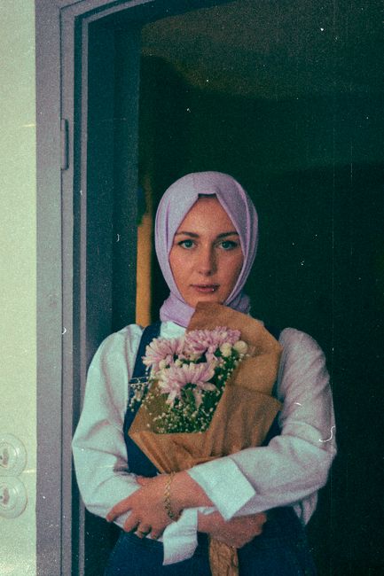 A woman in a hijab holding a bouquet of flowers, standing in a doorway.