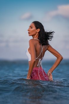 A woman in swimwear gracefully stands in the ocean with the sunset sky behind her.