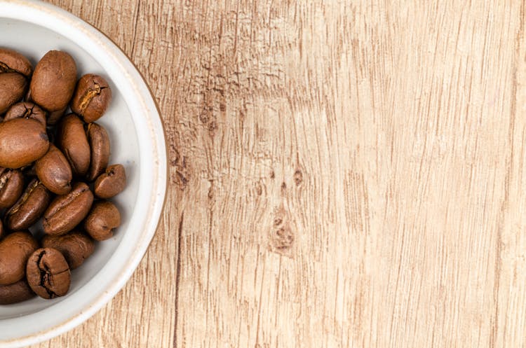 Coffee Beans On White Ceramic Bowl On Top Of Brown Wooden Surface