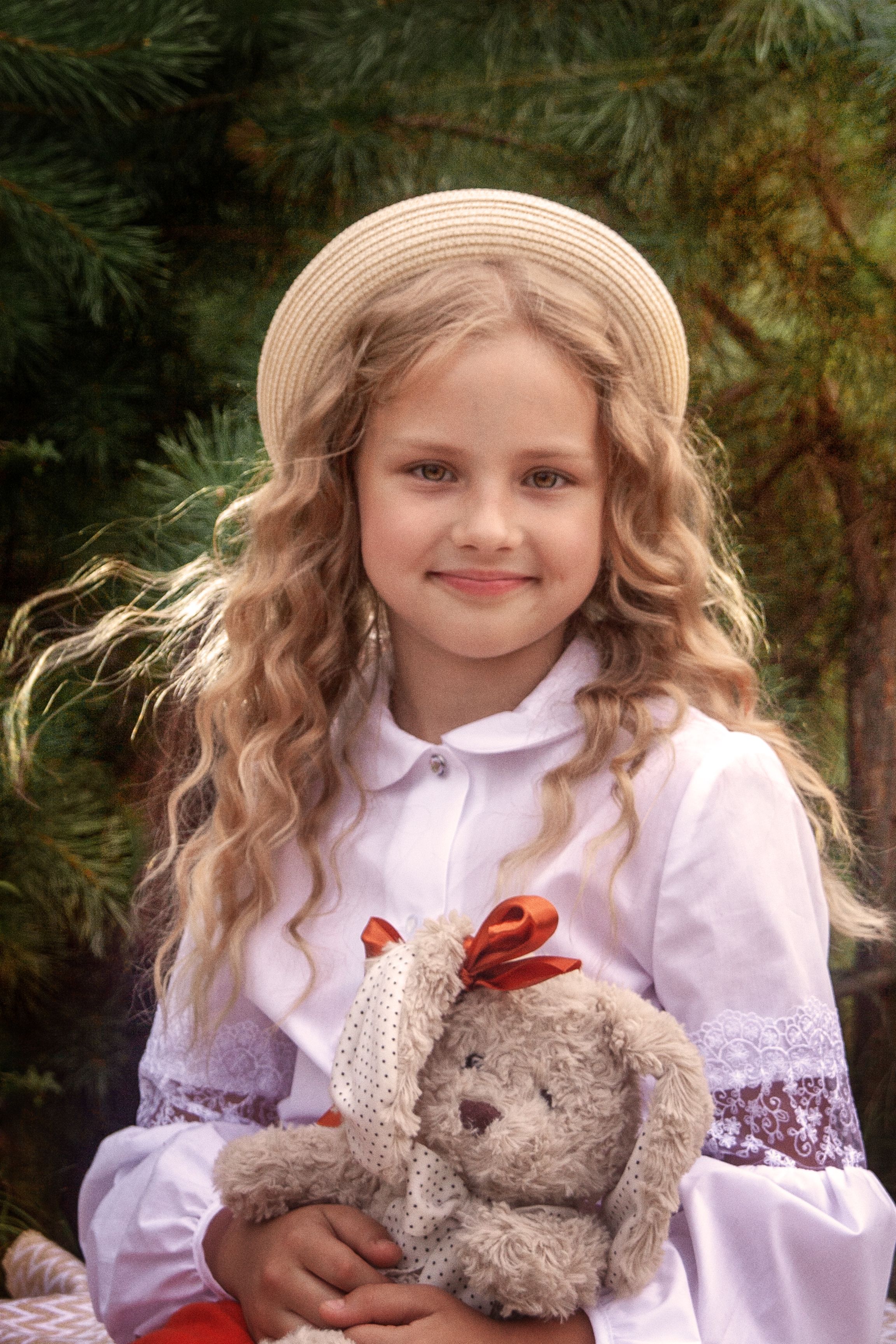 Charming young girl in a straw hat smiles while holding a cute teddy bear, surrounded by nature.