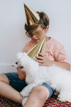 A child and fluffy dog wearing party hats and shutter shades indoors, celebrating together.