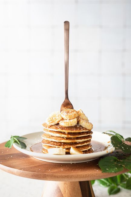 Stack of pancakes topped with banana slices, syrup, and herbs on a plate with a fork