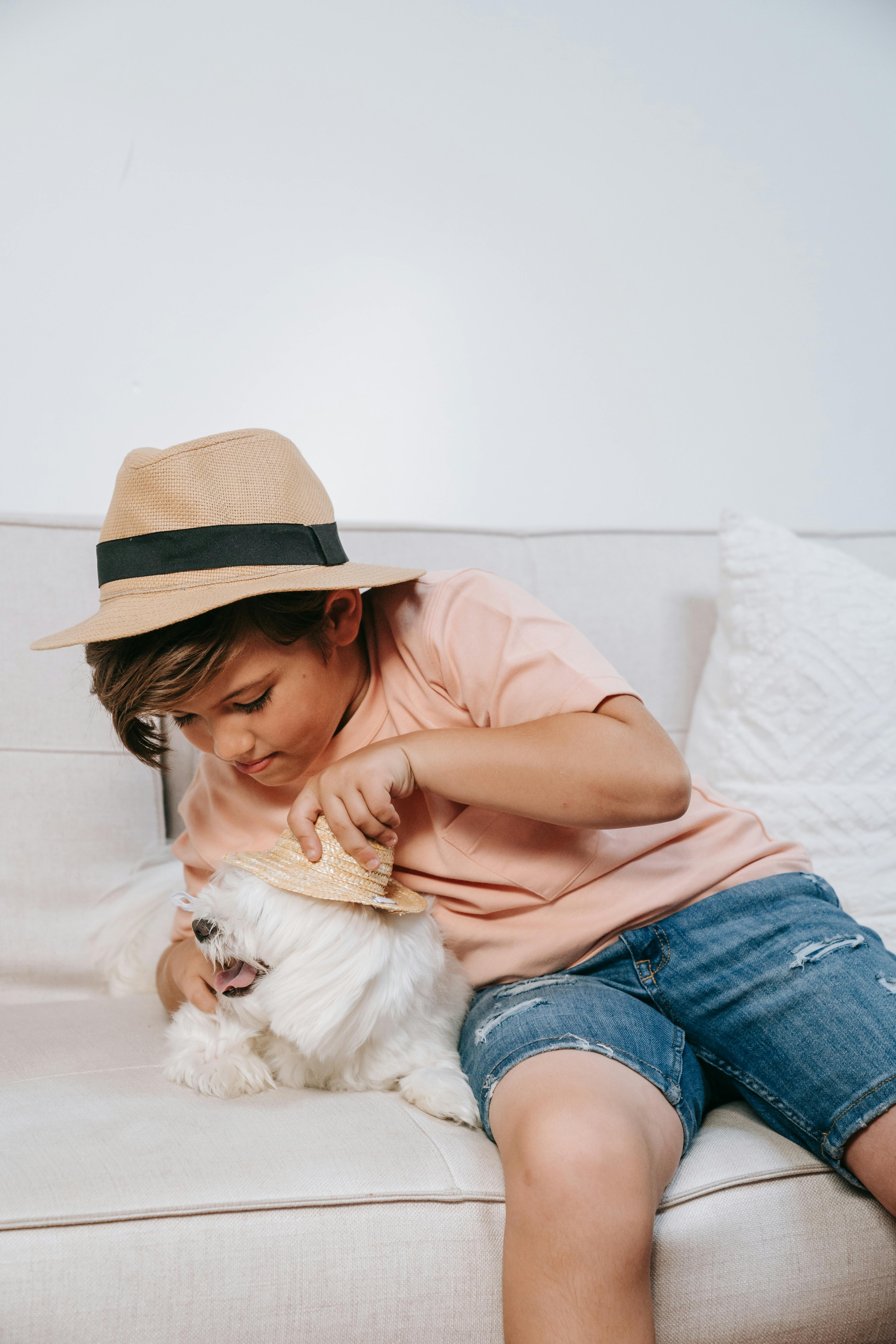 A child with a hat joyfully plays with a fluffy dog on a sofa, both wearing hats.