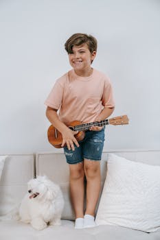 A cheerful child plays a ukulele while standing on a couch beside a fluffy white dog.