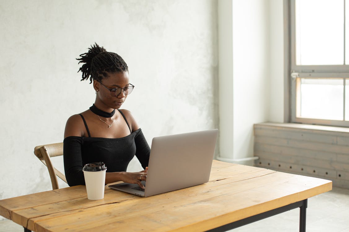 Concentrated African American female freelancer in casual clothes and eyeglasses sitting at table with cup of coffee near window and typing on netbook while looking at screen