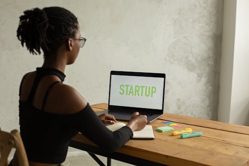 Back view of anonymous young African American businesswoman with bare shoulder and eyeglasses sitting at table with netbook with startup on screen and writing notes in copybook