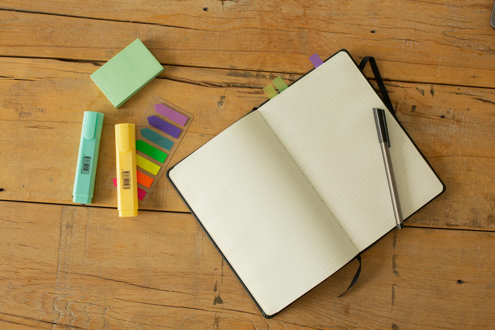 Top view of opened sketchbook with blank pages and pen with coloured markers and bookmarks arranged on wooden desk