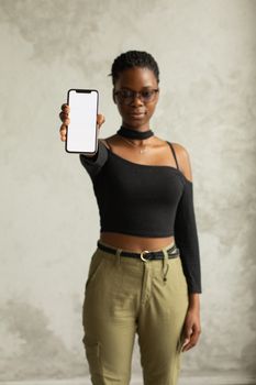 Unemotional young African American female in casual outfit and eyeglasses presenting cellphone with blank screen while looking at camera against white background - Fotografer Sidoarjo