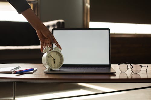 Free Anonymous black person touching alarm clock near opened netbook with white blank screen placed on table with notebook and pens Stock Photo