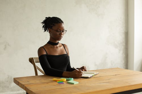 Concentrated young African American female in black outfit and eyeglasses sitting at wooden table and writing information with pencil and colourful markers against white wall