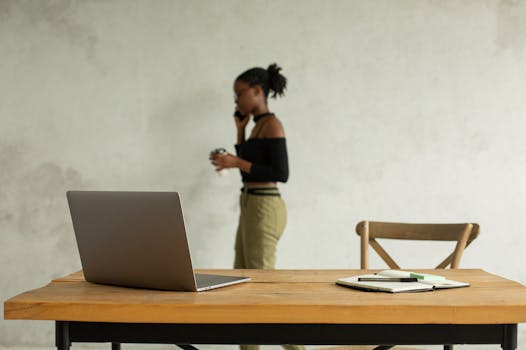 A laptop and notebook on a wooden table with a woman talking on the phone in the background.