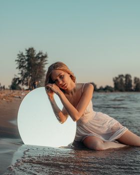 A woman in a white dress poses with a mirror on a tranquil beach at sunset.