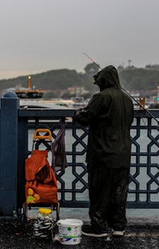 A solitary fisherman in a raincoat stands by the harbor fishing in the rain.