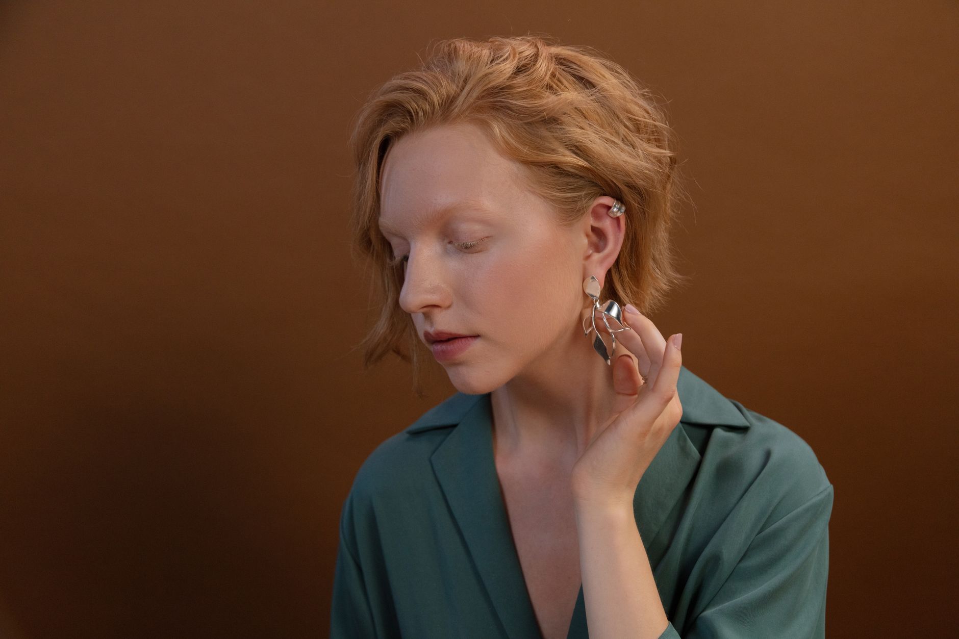Close-up of a woman with ginger hair and elegant earrings, posing against a brown background.