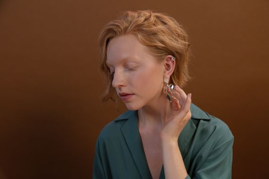 Close-up of a woman with ginger hair and elegant earrings, posing against a brown background.