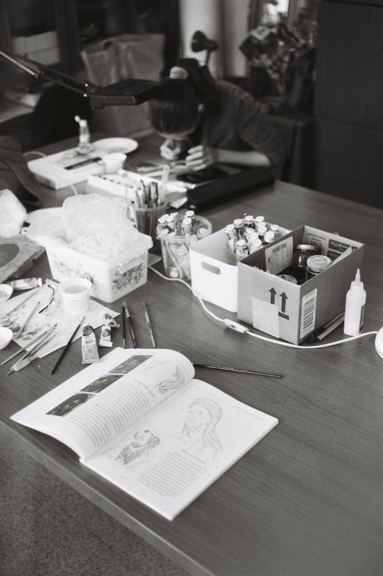 Black And White Photograph Of A Desk With Painting Materials And A Catalog