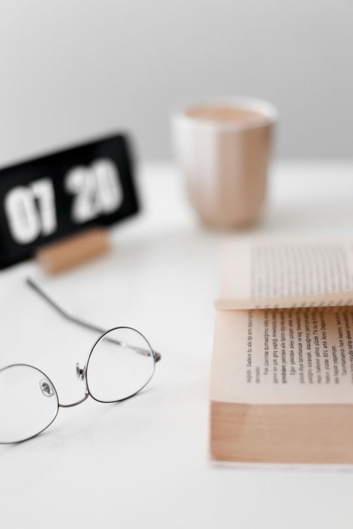 Free Close-up of an open book, round eyeglasses, a cup, and a clock displaying 07:20 on a white background. Stock Photo