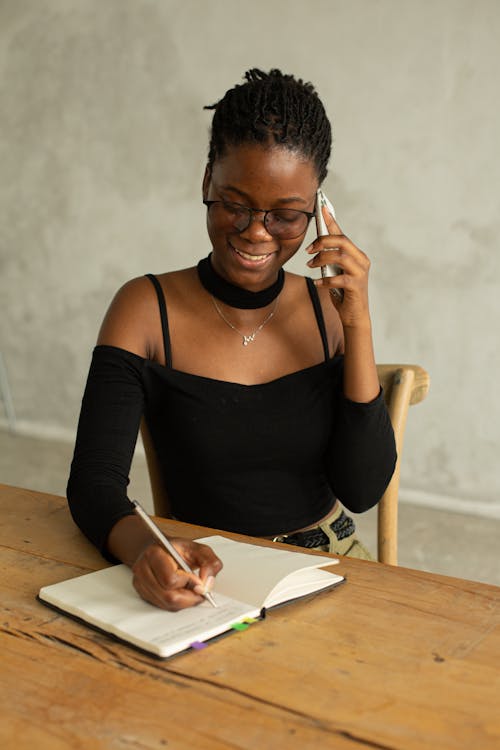 Cheerful young African American female sitting at table and taking notes in notepad while having conversation on mobile phone in light room